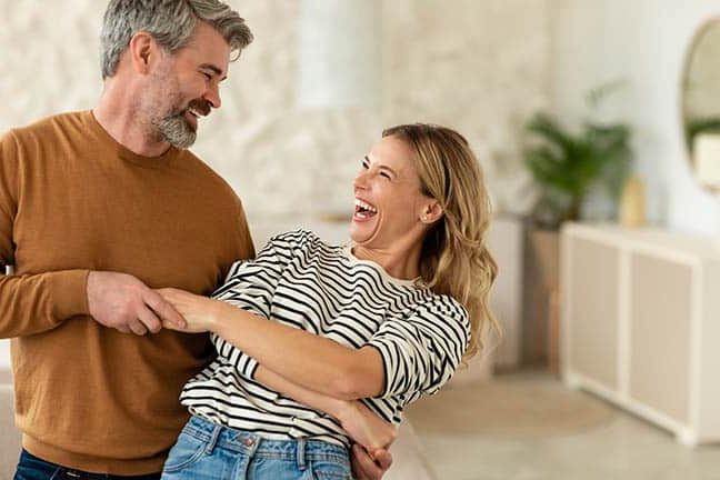 Joyful Middle Aged Spouses Dancing In Living Room At Home woman with dental implants dancing with her husband