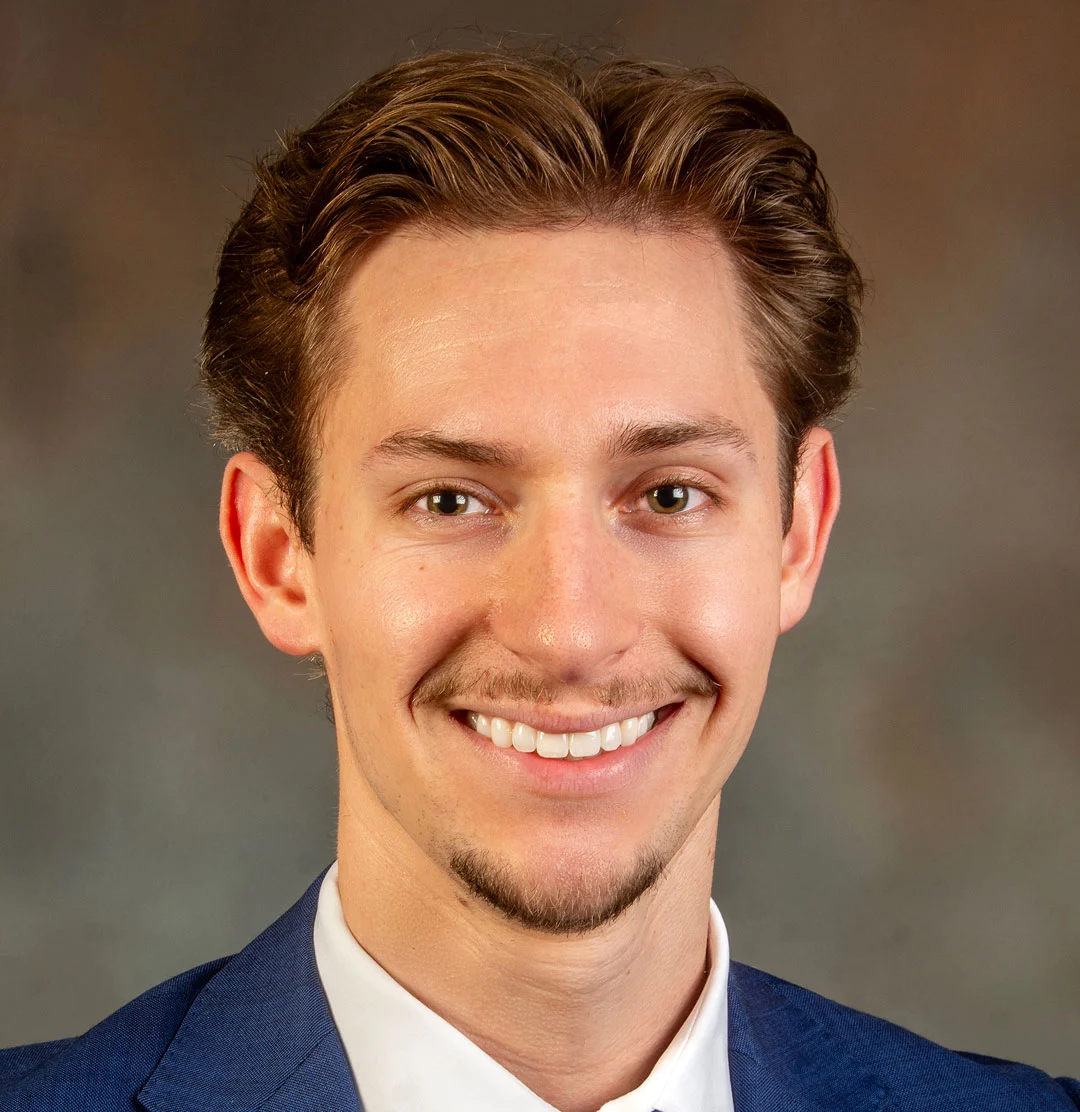 A young man with light brown hair, a trimmed goatee, and a blue suit smiles at the camera in front of a neutral brown background.